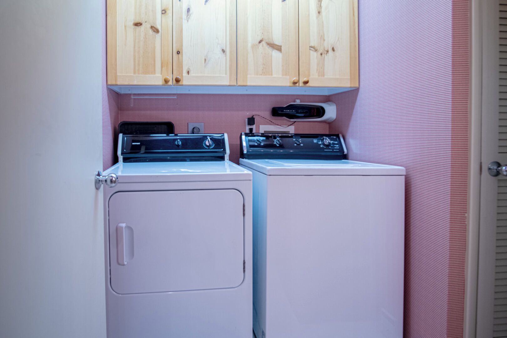 A laundry room with two washers and dryers, wooden cabinets above, and pink checkered walls.