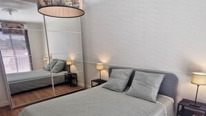 Et moderne soveværelse med et stort spejl, en seng med gråt sengetøj, to sengeborde med lamper og trægulv.