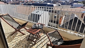 En balkon med udsigt over bybilledet, med flisegulv, træmøbler og gelænder i hvid.