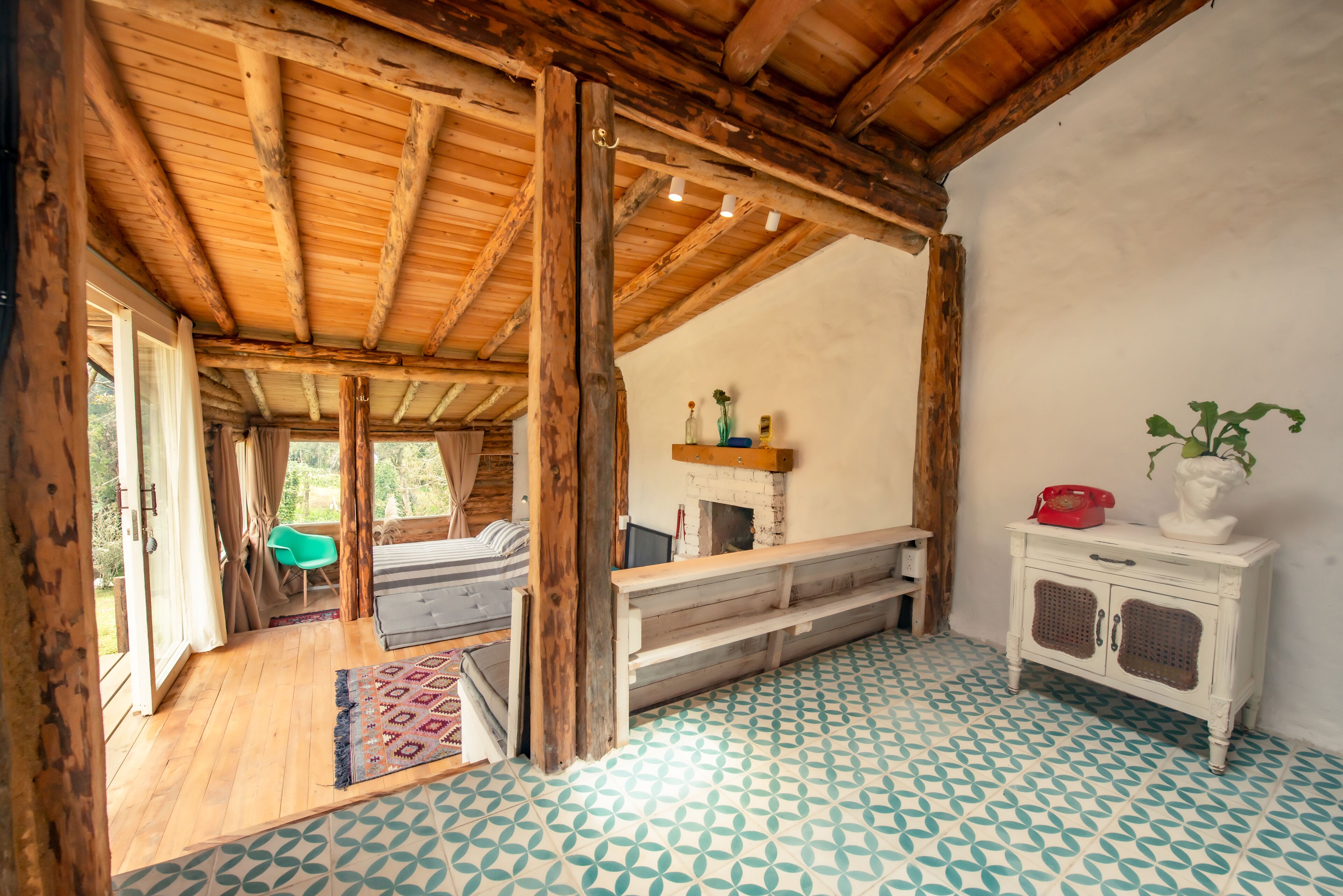 A rustic room with a wooden ceiling, a patterned floor, a bench, a small table with a vase, and a cabinet with a telephone.