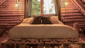 Quarto de cabana de toras aconchegante, com cama, almofadas e vista para árvores pela janela.