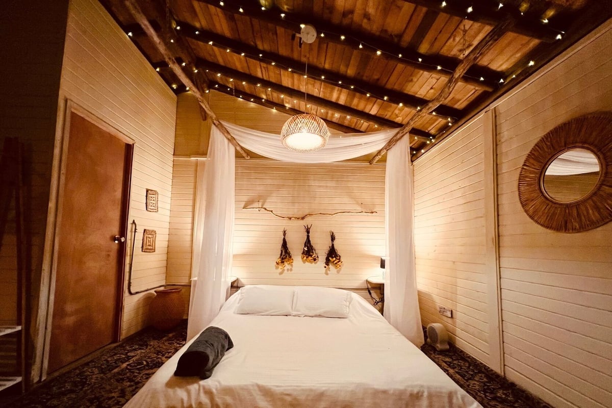 Een knus slaapkamer met een houten plafond, een bed met witte lakens, een hanglamp en een ronde spiegel aan de muur.