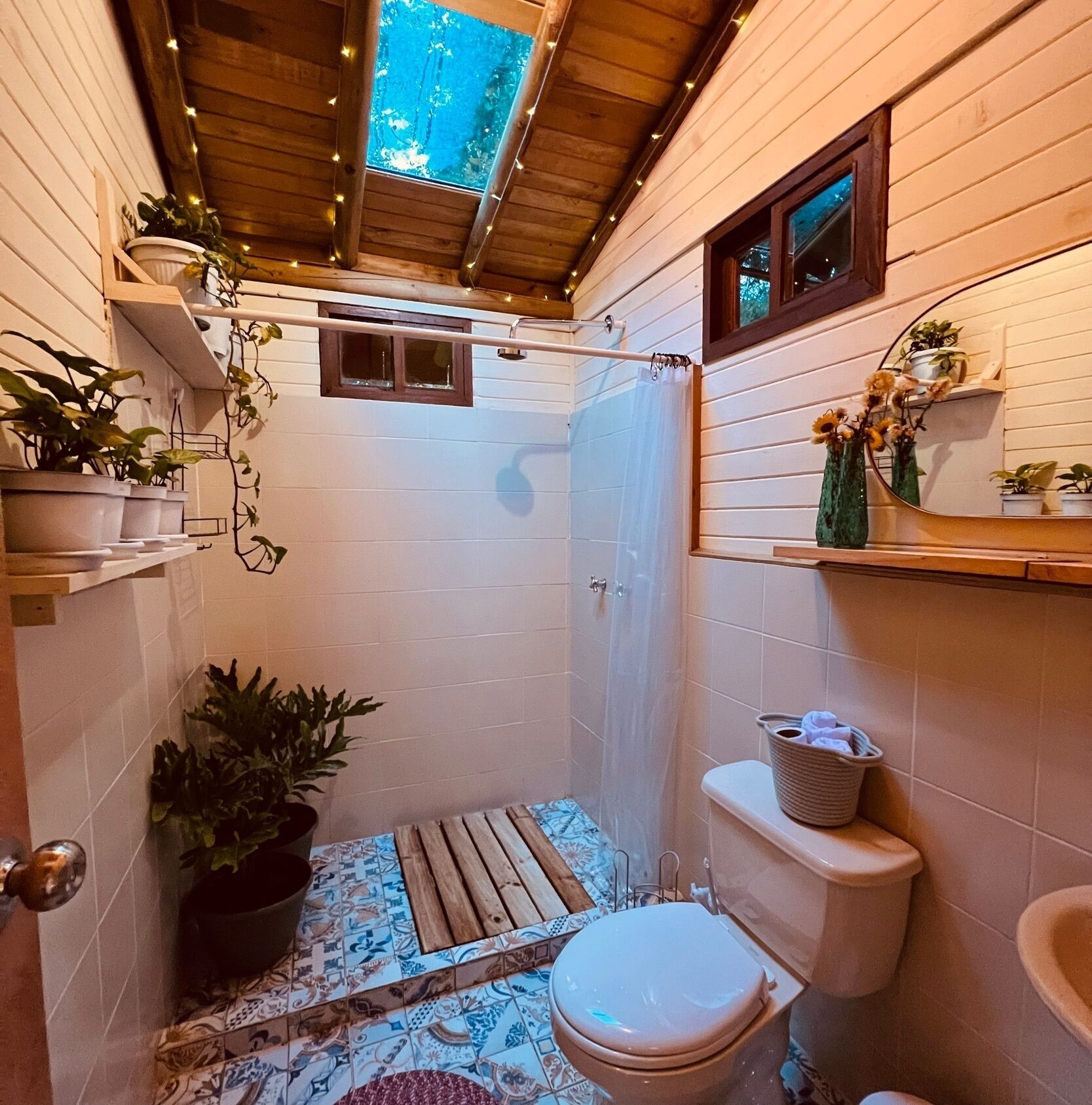 A small, narrow bathroom with a shower, toilet, and a small window. There are potted plants and a wooden shelf with toiletries.
