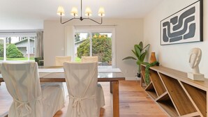 Un comedor con una mesa de madera, sillas blancas y una araña. Se observa un ventanal con vistas a zonas verdes.