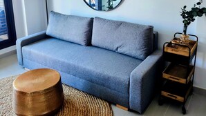 A modern living room with a grey sofa, a round mirror, a woven rug, and a wooden side table with a plant. - Apartamento Moderno y Confortable con Piscina Comunitaria (Verger)