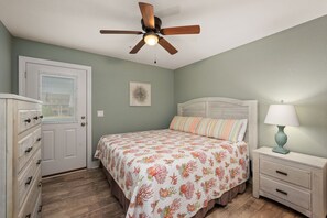 Une chambre à coucher comprenant un lit, une table de chevet, une commode, un ventilateur de plafond et une porte avec des stores.