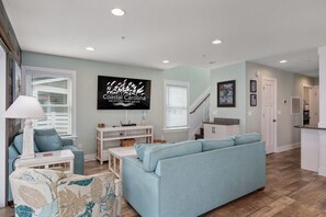 Una sala de estar moderna con un sofá azul claro, un sillón estampado y una mesa de centro blanca. Un televisor de pantalla plana está montado en la pared, y hay una escalera que lleva a otro nivel.