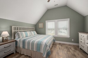 Quarto com teto inclinado, cama com colcha listrada, mesa de cabeceira com luminária, cômoda e janela com persianas.
