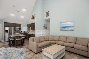 A modern kitchen with a built-in oven, a dining area with a table and chairs, a large L-shaped sectional sofa, and a painting of a beach scene. - Haven on the Banks: Semi-Oceanfront, Community Pool, Perfect for Wedding Parties (Nags Head)
