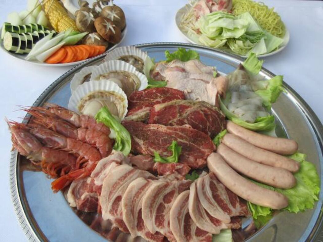 Eine Platte mit verschiedenen Fleisch- und Fischsorten, darunter Scheiben Rindfleisch, Würstchen und Garnelen, zusammen mit Gemüse wie Salat und Mais.