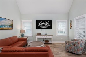 A living room with a sofa, a round table, a TV mounted on the wall, and a window with blinds.