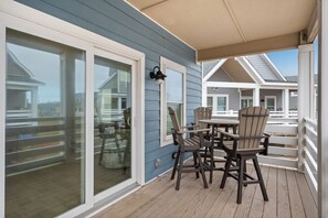 A covered outdoor area with a table and chairs, a glass door leading to a deck, and a building with blue siding.
