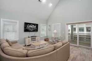 A living room with a large beige sectional sofa, a flat-screen TV mounted on the wall, a small white cabinet, and a wicker basket on the floor.