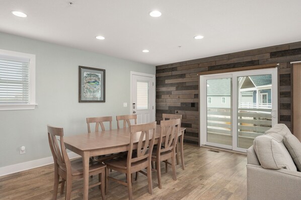 Un coin repas avec une table et des chaises en bois, une porte avec une fenêtre, et une vue sur une maison à travers une porte-fenêtre coulissante.