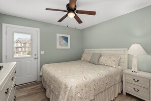 Quarto com cama, mesa de cabeceira, cômoda, ventilador de teto e janela com vista para edifícios.
