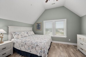 Une chambre avec un plafond en pente, un lit recouvert d’une couvre-lit à motifs, une table de chevet avec une lampe et une fenêtre avec des stores.