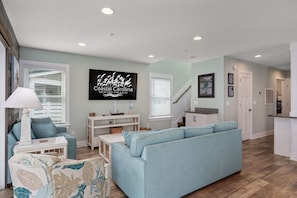 A modern living room with a light blue sofa, a patterned armchair, and a white coffee table. A flat-screen TV is mounted on the wall, and there is a staircase leading to another level. - Currituck Cottage: Semi-Oceanfront, Private Hot Tub, Community Pool Access! (Nags Head)