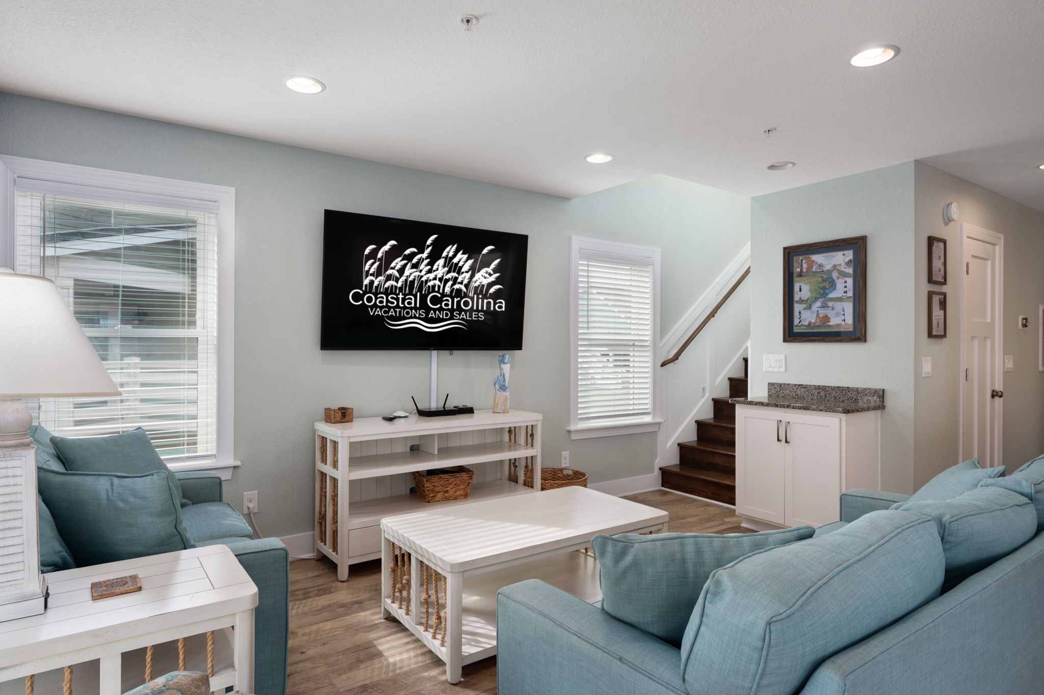 A living room with teal sofas, a white coffee table, and a TV mounted on the wall displaying 'Coastal Carolina Vacations and Sales'.