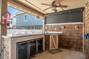 A covered outdoor kitchen area with a ceiling fan, built-in cabinets, and a view of neighboring houses. - Currituck Cottage: Semi-Oceanfront, Private Hot Tub, Community Pool Access! (Nags Head)