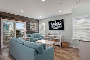 A modern living room with a large TV, light blue sofa, and a wooden accent wall. - Currituck Cottage: Semi-Oceanfront, Private Hot Tub, Community Pool Access! (Nags Head)