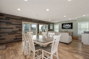 A modern living room with a dining area, wooden flooring, and a large TV screen displaying 'Coastal Carolina'.