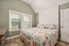 A bedroom with a bed, two windows with blinds, a dresser, and a door.