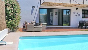 Une piscine avec un espace patio, du mobilier d’extérieur et un bâtiment doté de grandes fenêtres et d’une pergola.