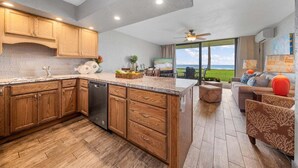 A modern kitchen with wooden cabinets, a dishwasher, and a large island. There is a dining area with a sofa and a view of the outdoors.