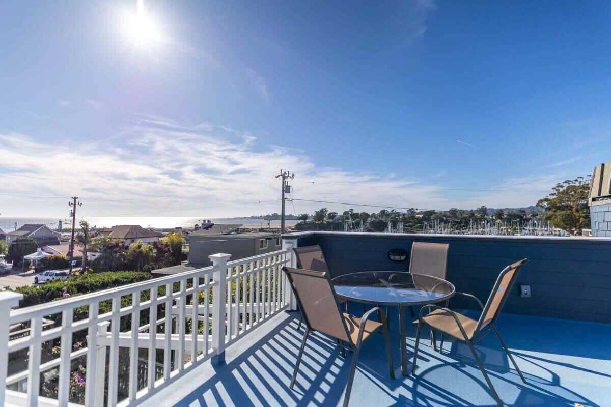 En balkon med bord og stole, udsigt over et kystområde med huse og en klar himmel.