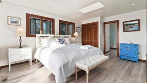 A bedroom with a large bed, a bench, a nightstand, a dresser, and a door leading to another room.