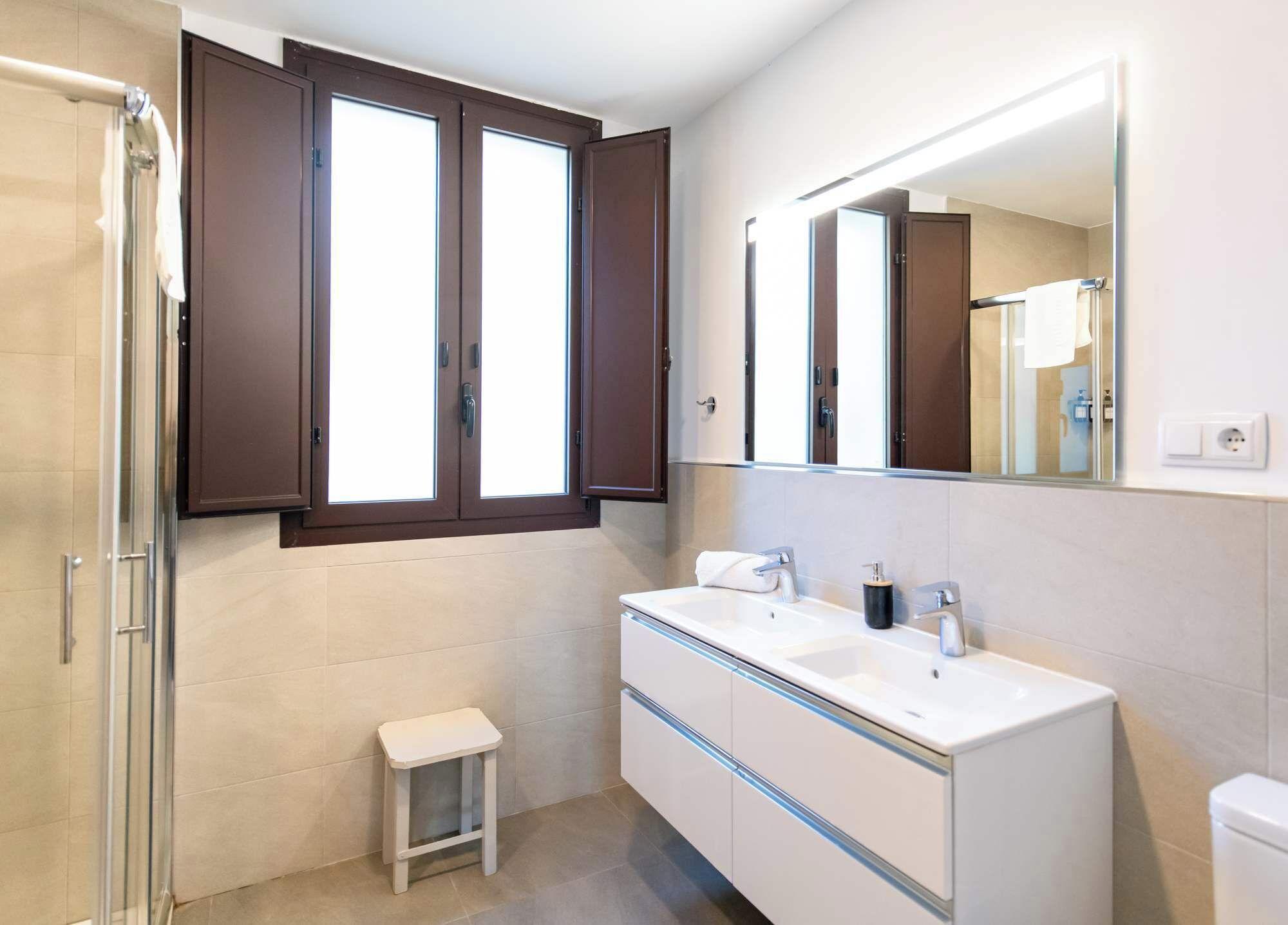 Un baño moderno con tocador doble, un taburete pequeño, mampara de ducha de vidrio y un espejo grande.