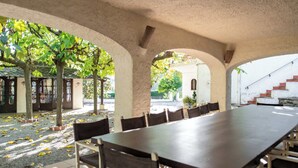 An outdoor dining area with a long table and chairs, surrounded by arches and trees. - Unique 16th-Century Masia: Pool & Private Events (Barcelona)