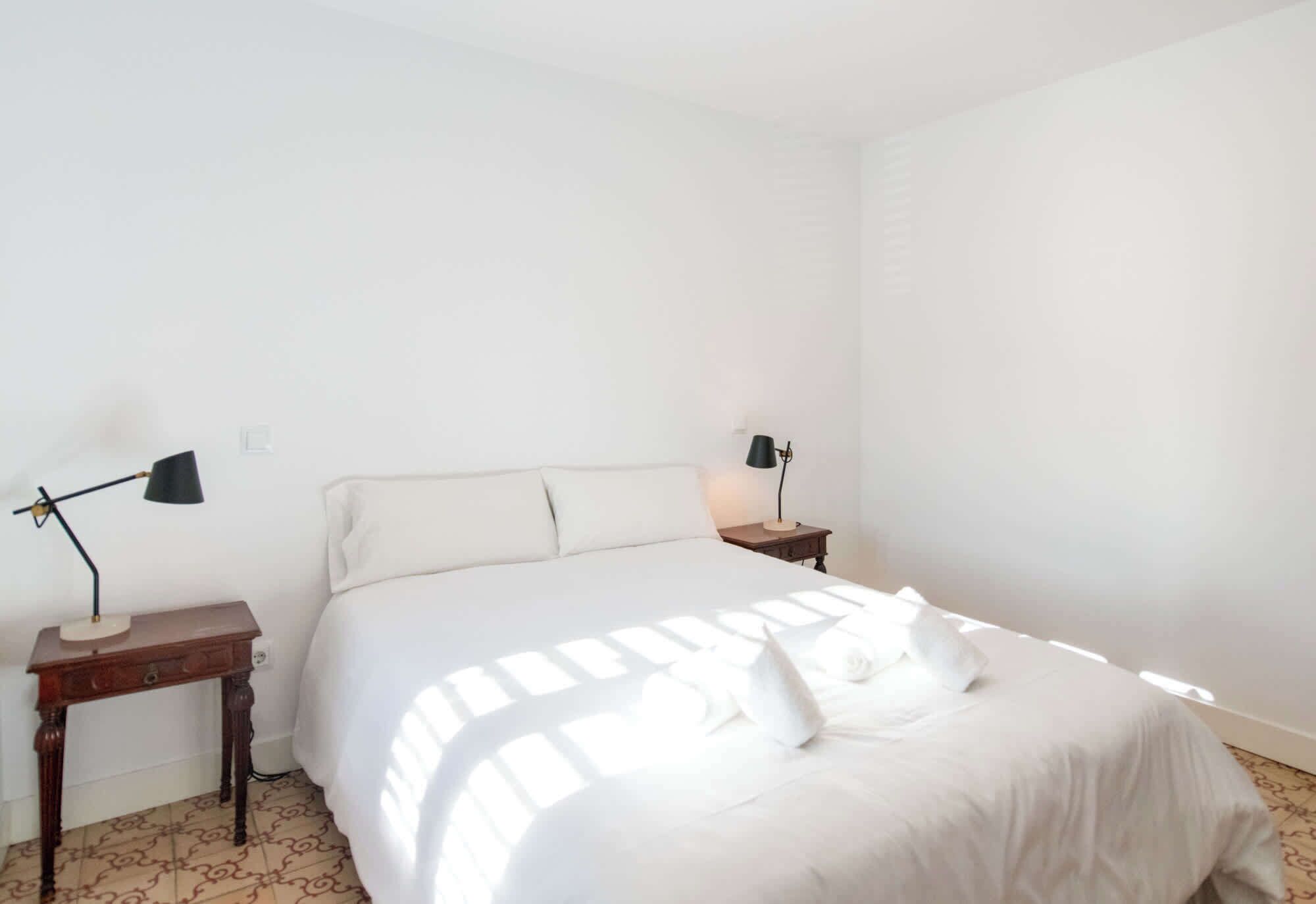 Un lit bien fait, avec du linge de lit blanc et des oreillers, deux tables de chevet avec des lampes et un mur blanc sobre.
