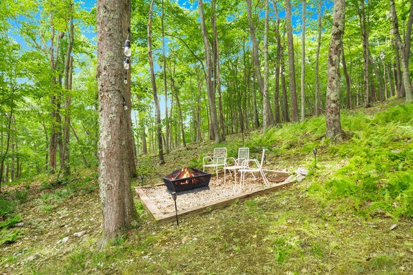 A forest campsite with a fire pit, two white chairs, and a metal table.
