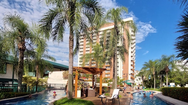 A swimming pool with palm trees and lounge chairs, surrounded by a building with balconies. - Morada Stay - Flat Barra First (Rio de Janeiro)