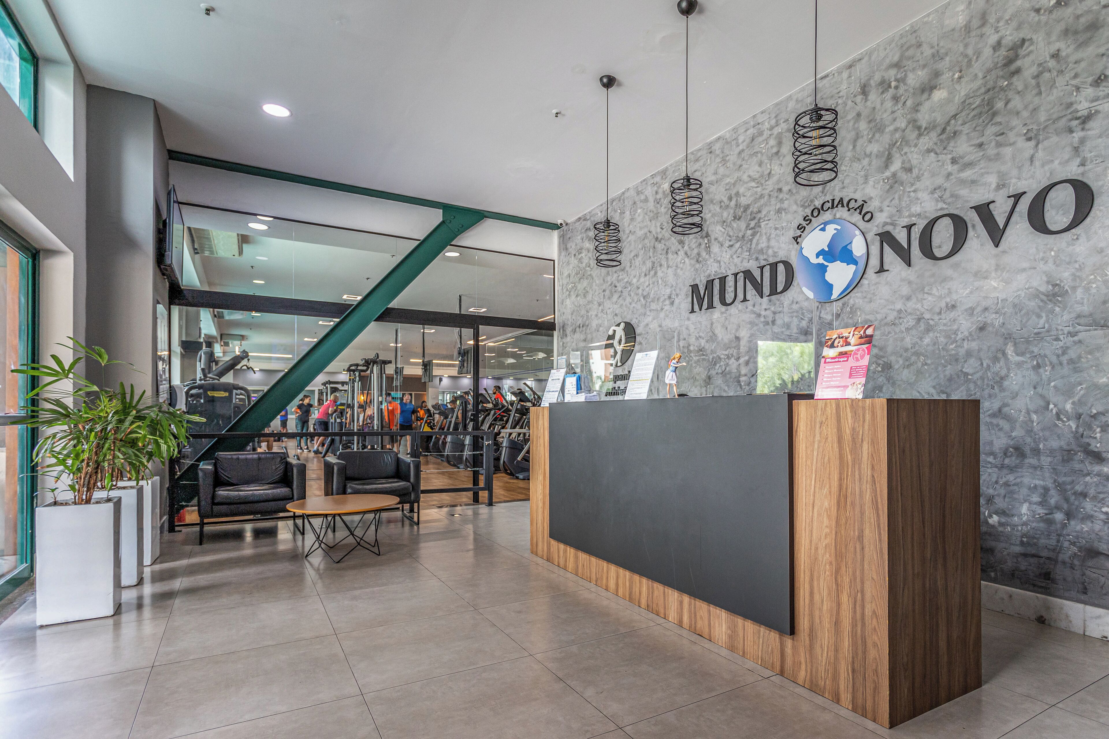 A reception desk with a wooden finish and a black panel, a grey wall with the text "MUND NOVO" and a globe logo, and a staircase leading to upper floors.