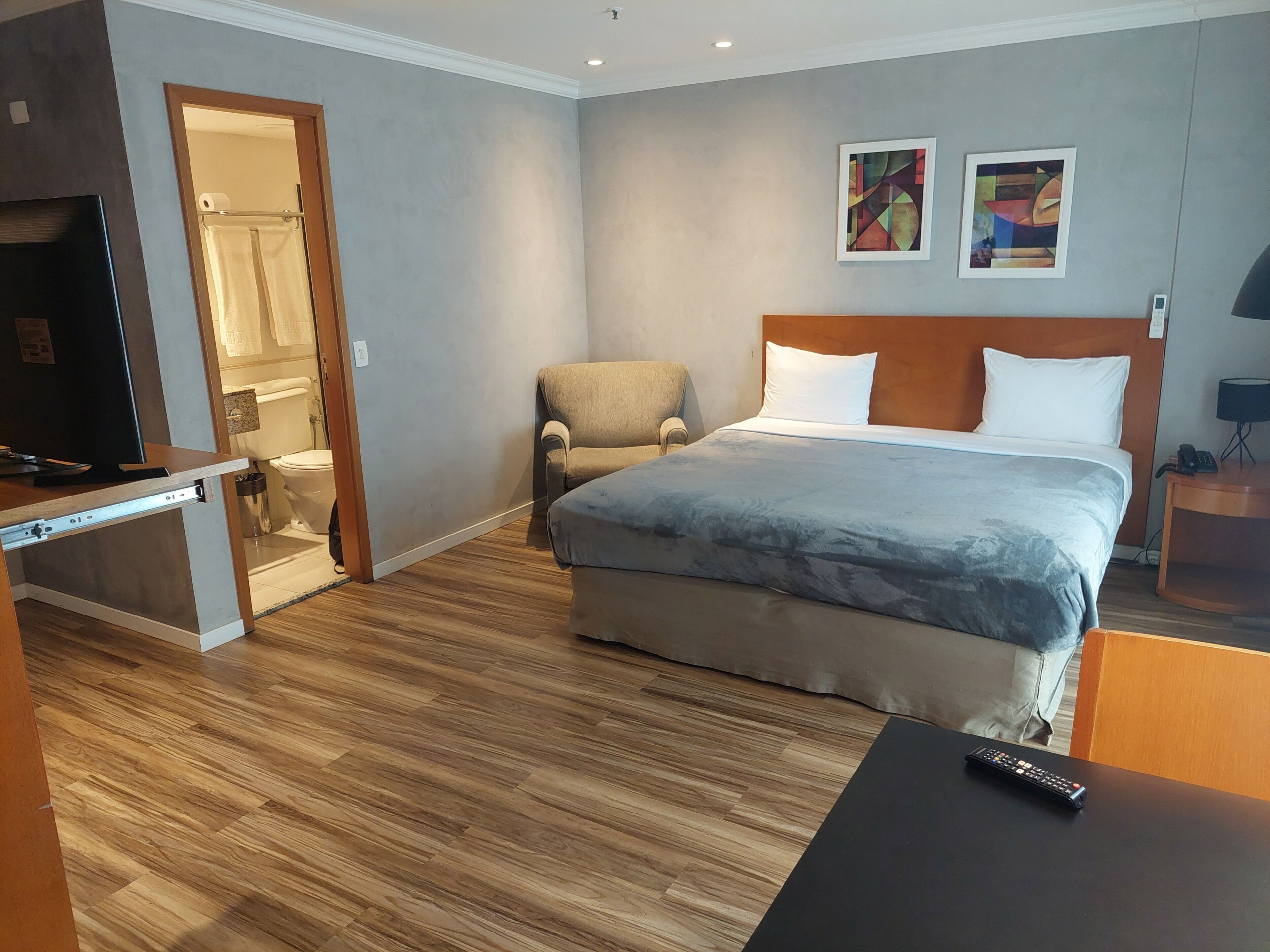 A hotel room with a bed, a chair, a desk with a TV, and a bathroom visible through an open door.