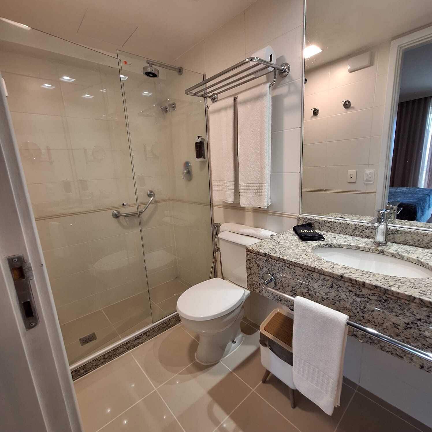Un baño moderno con ducha envuelta en vidrio, inodoro blanco, lavamanos con encimera de granito y un espejo sobre el lavabo.