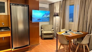 A hotel room with a wooden floor, a minibar, a flat-screen TV, a dining area with a table and chairs, and a view of the outdoors through a window. - Morada Stay - Flat Barra First (Rio de Janeiro)