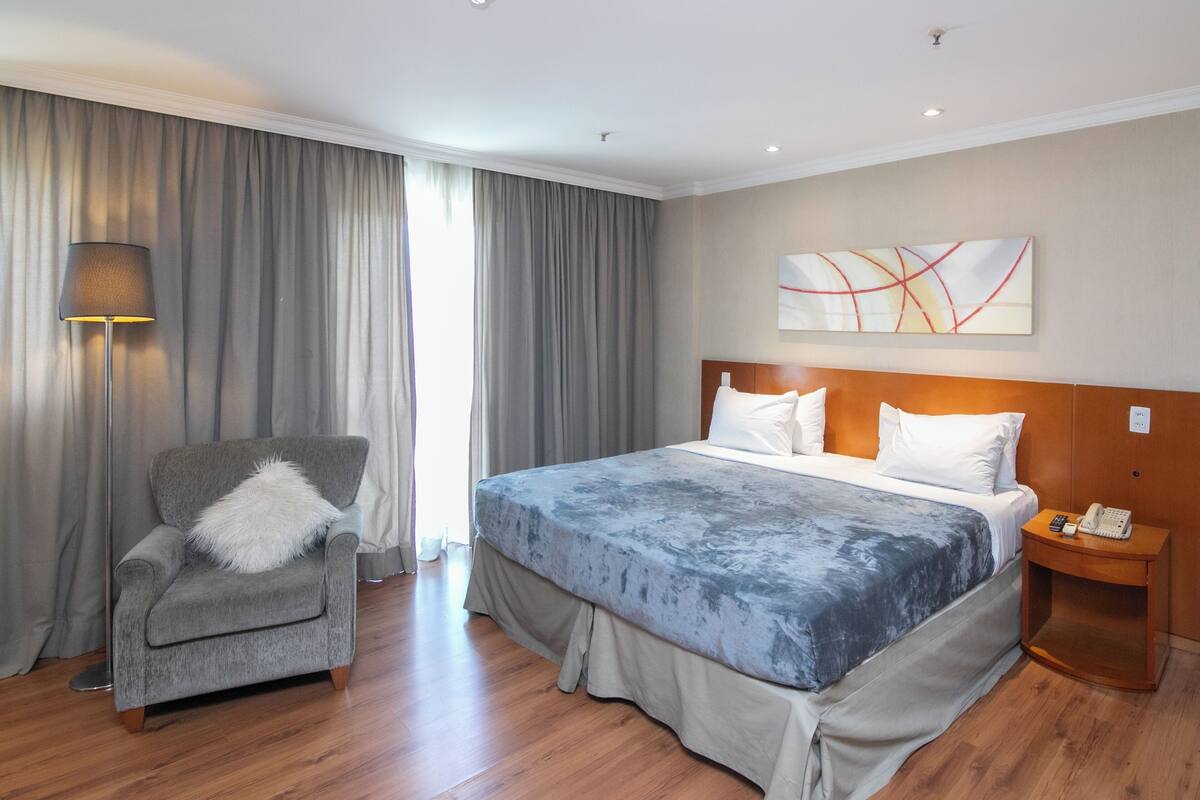 A hotel room with a bed, a chair, a nightstand, a lamp, and a painting on the wall.