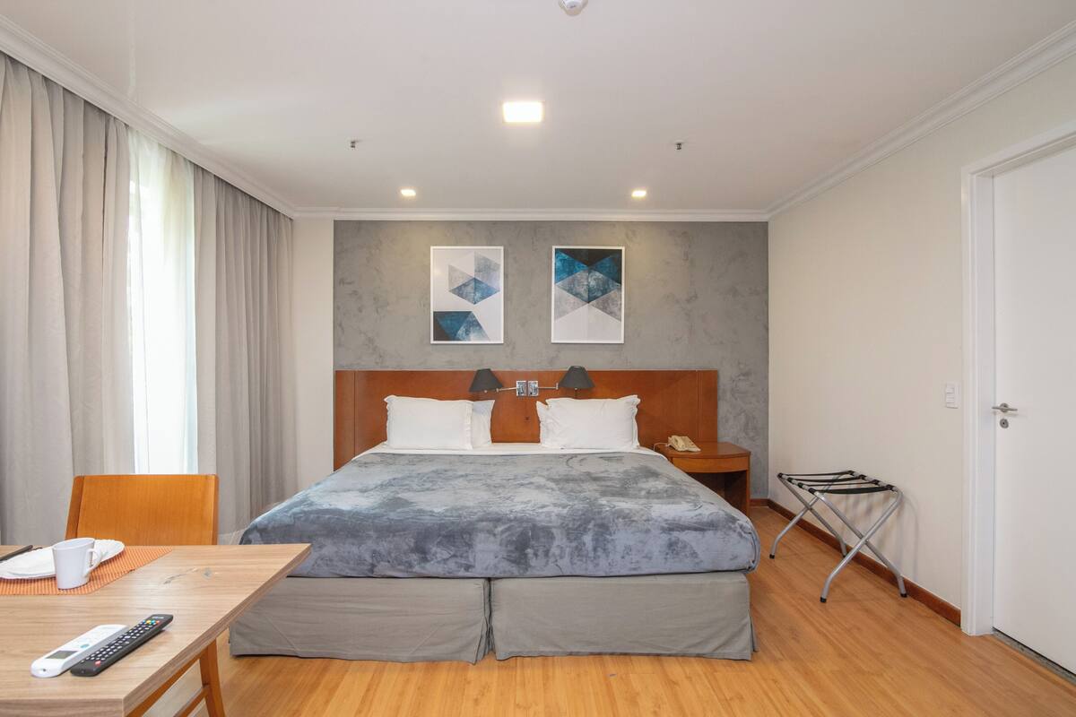 A hotel room with a bed, a desk with a remote control, a nightstand, and a wall-mounted lamp.