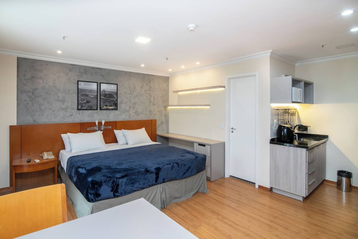 A hotel room with a bed, bedside table, desk, kitchenette, and a bathroom.
