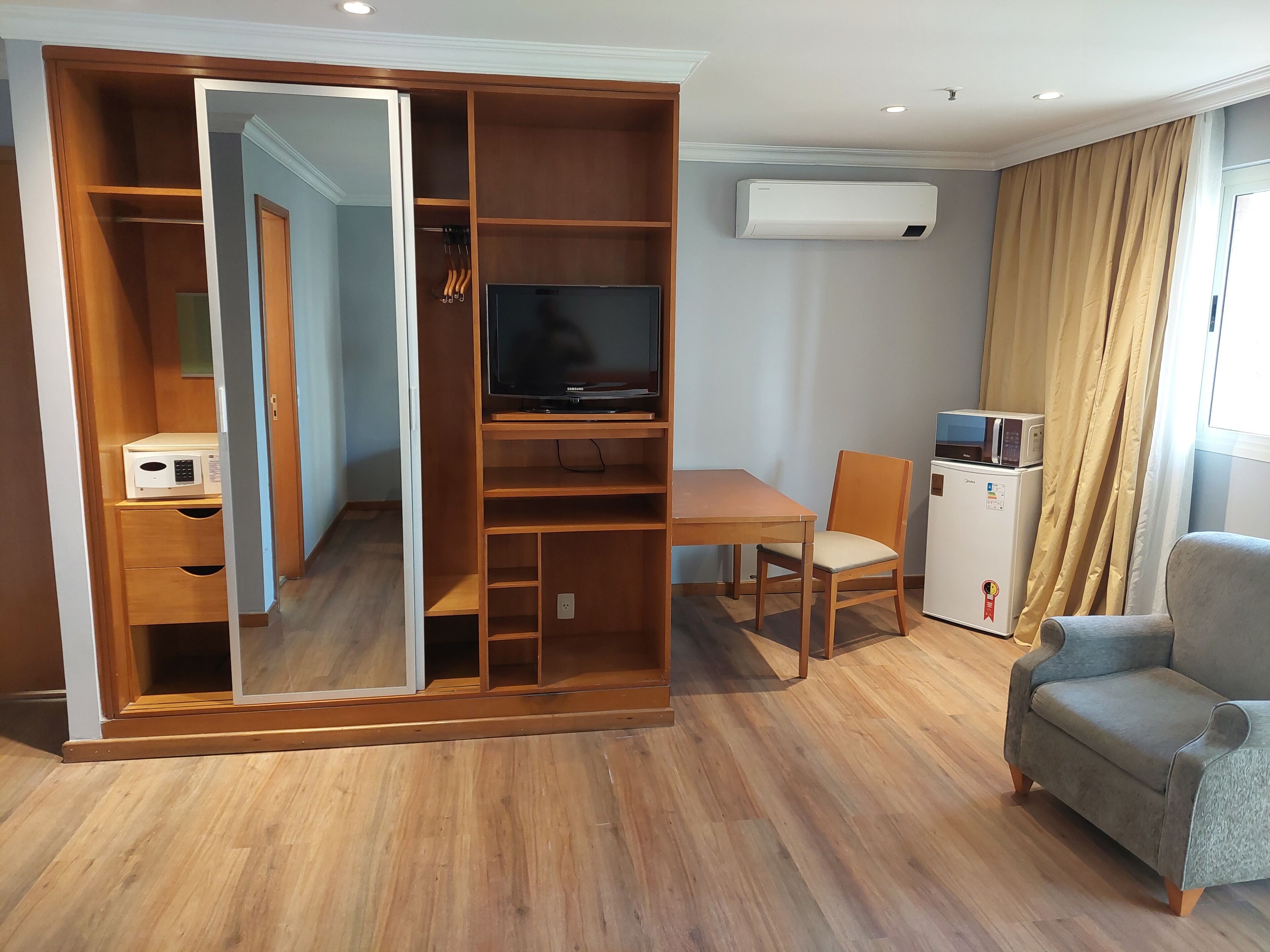 Un salon moderne comprenant un meuble de rangement en bois pour l’équipement audiovisuel, un téléviseur à écran plat, une table à manger avec des chaises, un canapé, un réfrigérateur et un appareil de climatisation.