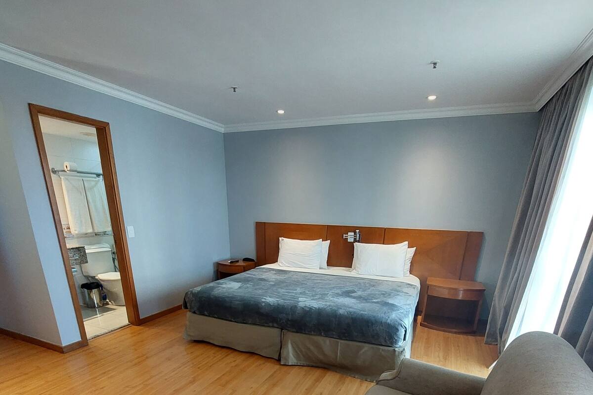 A hotel room with a bed, a chair, a nightstand, and a view of the outside through a window.