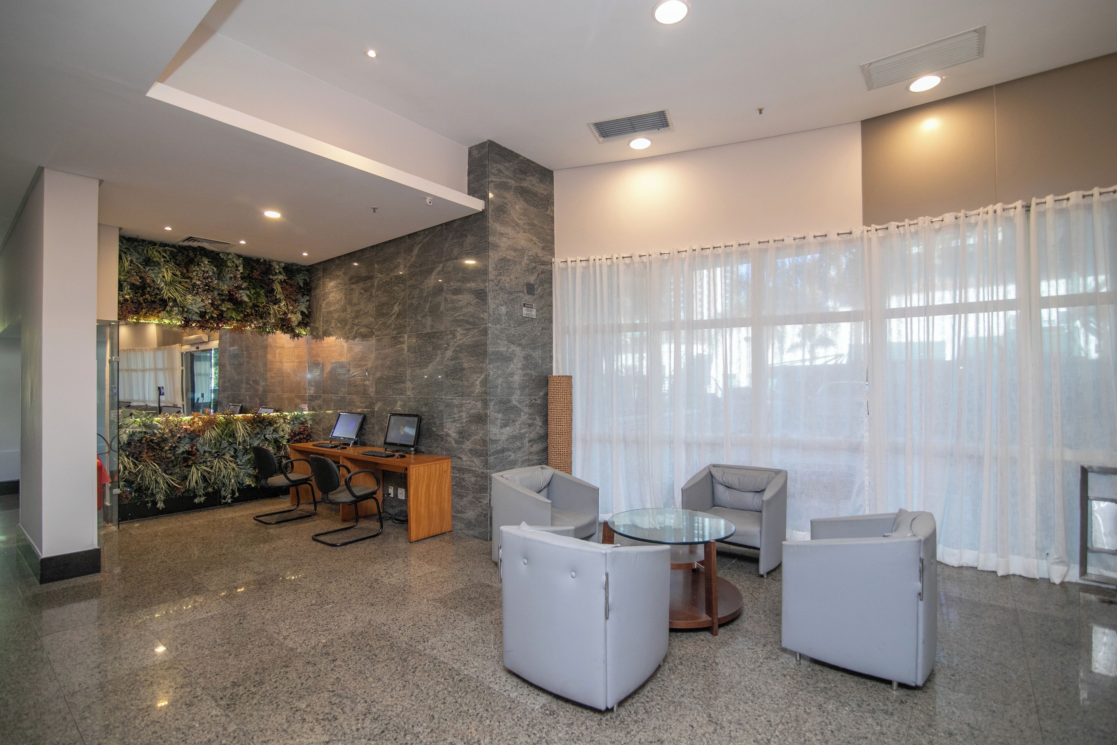 A modern lobby with a glass coffee table, grey sofas, a desk with computers, and a decorative wall with plants.