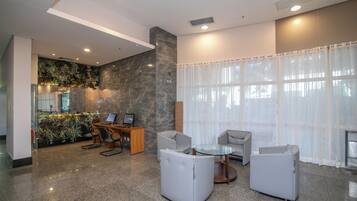 A modern lobby with a glass coffee table, grey sofas, a desk with computers, and a decorative wall with plants.