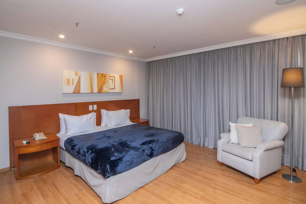 A hotel room with a bed, a nightstand, a chair, a lamp, and a curtain.