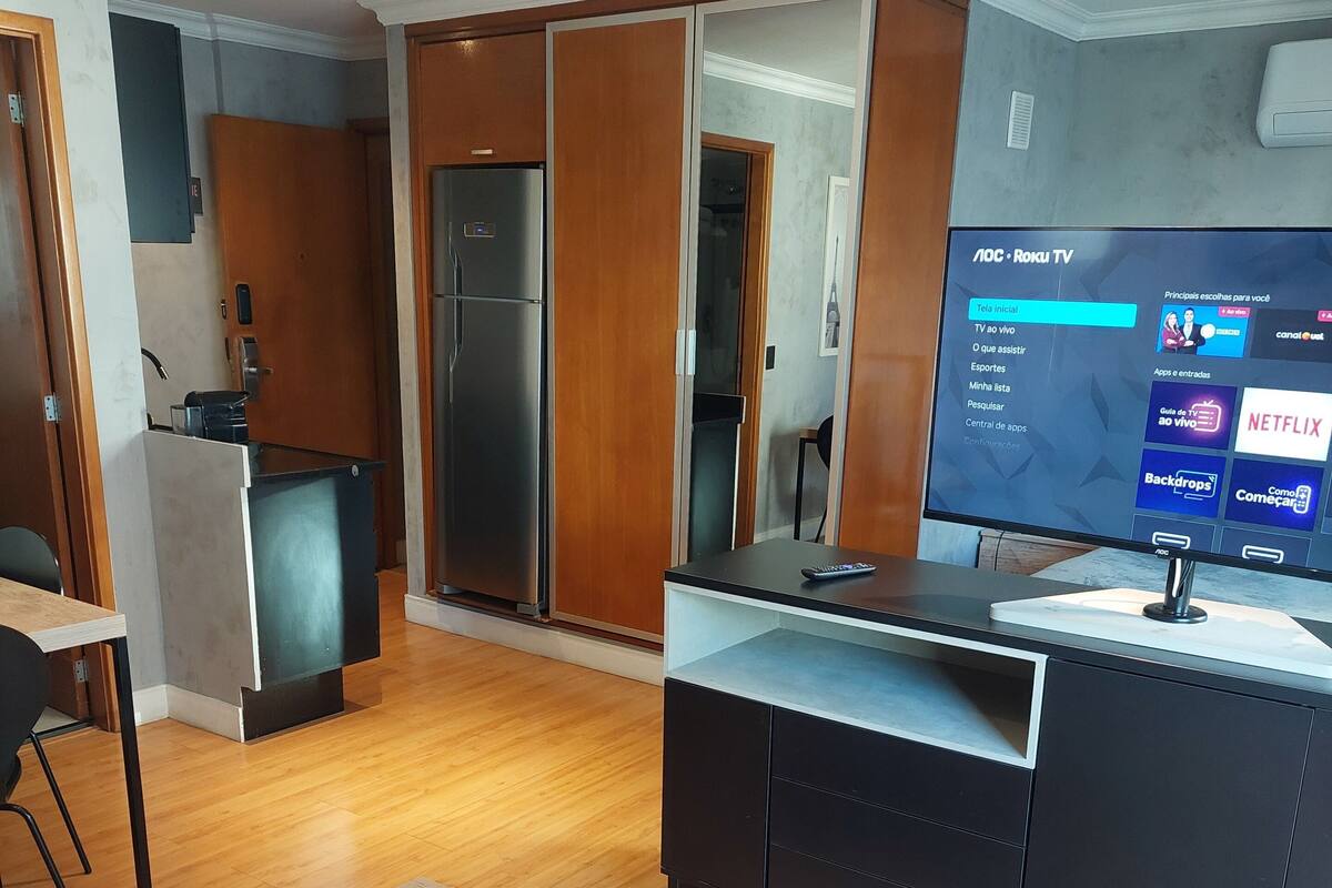 A modern living room with a wooden floor, a TV, a kitchen area with a refrigerator, and a dining table.