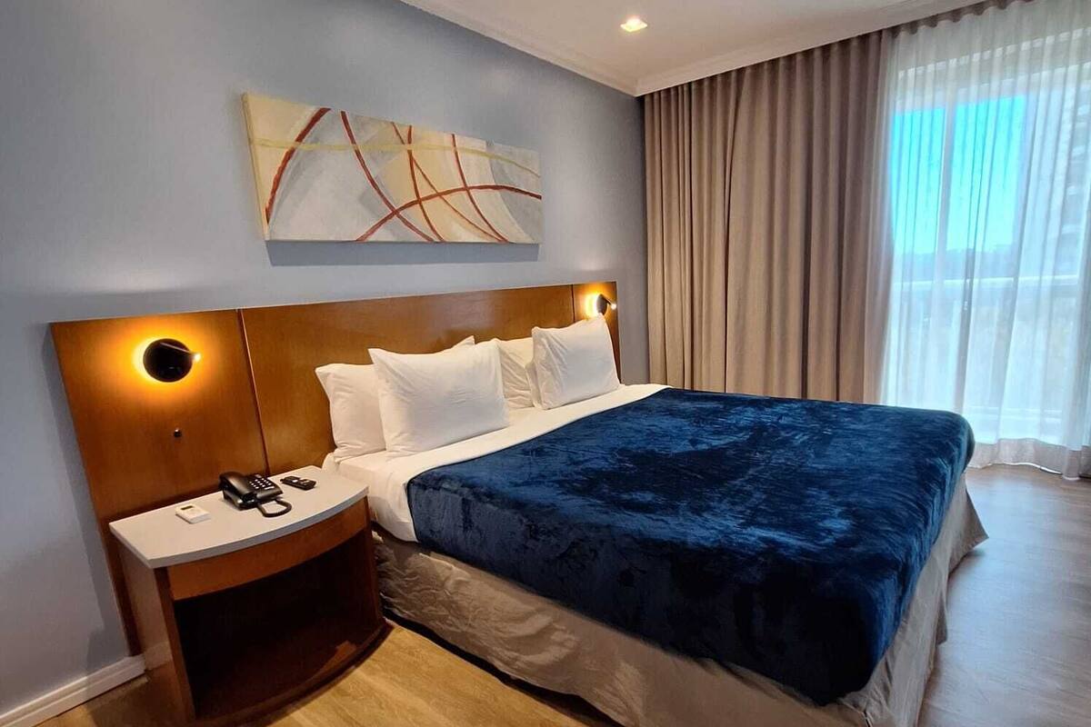 A hotel room with a large bed, a nightstand with a phone, a painting on the wall, and a window with curtains.
