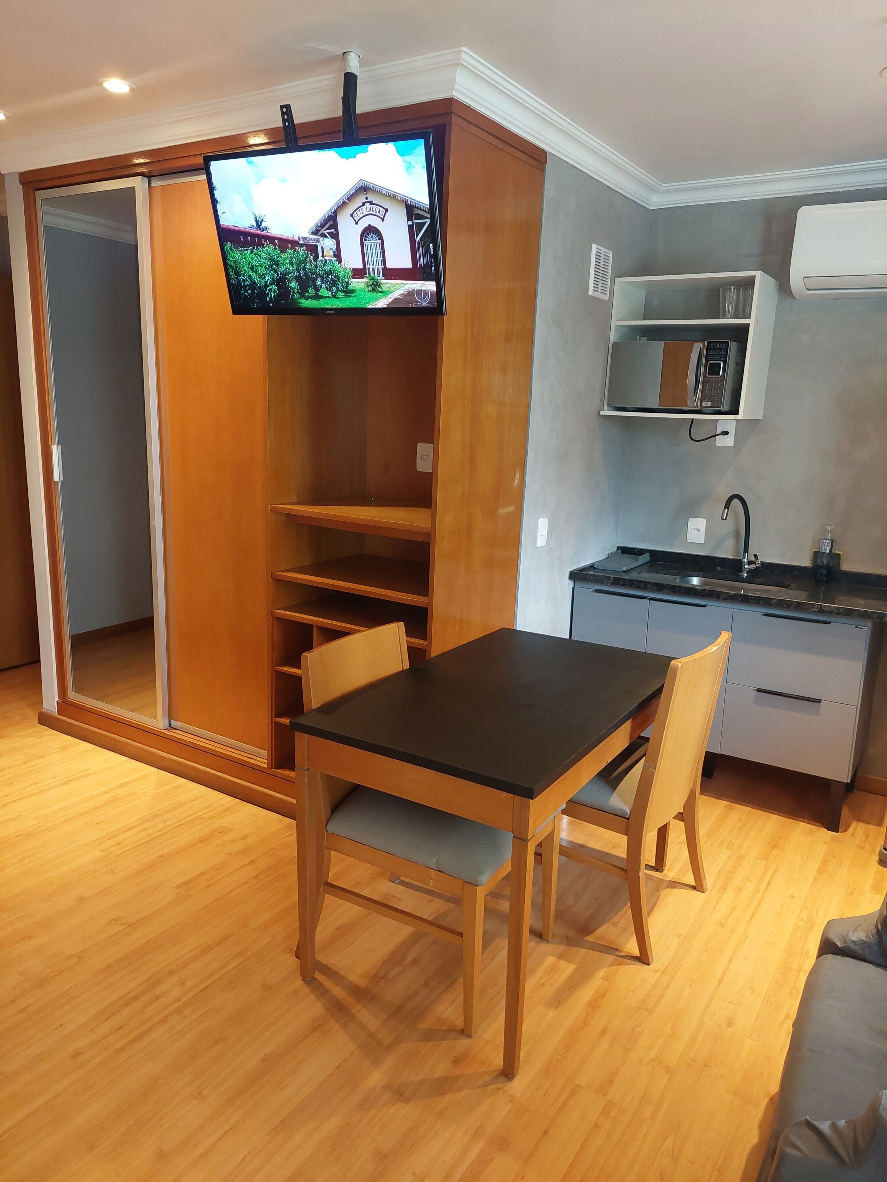 A compact living space with a wooden dining table, a television mounted on a cabinet, a kitchenette with a microwave, and a sliding door leading to another room.
